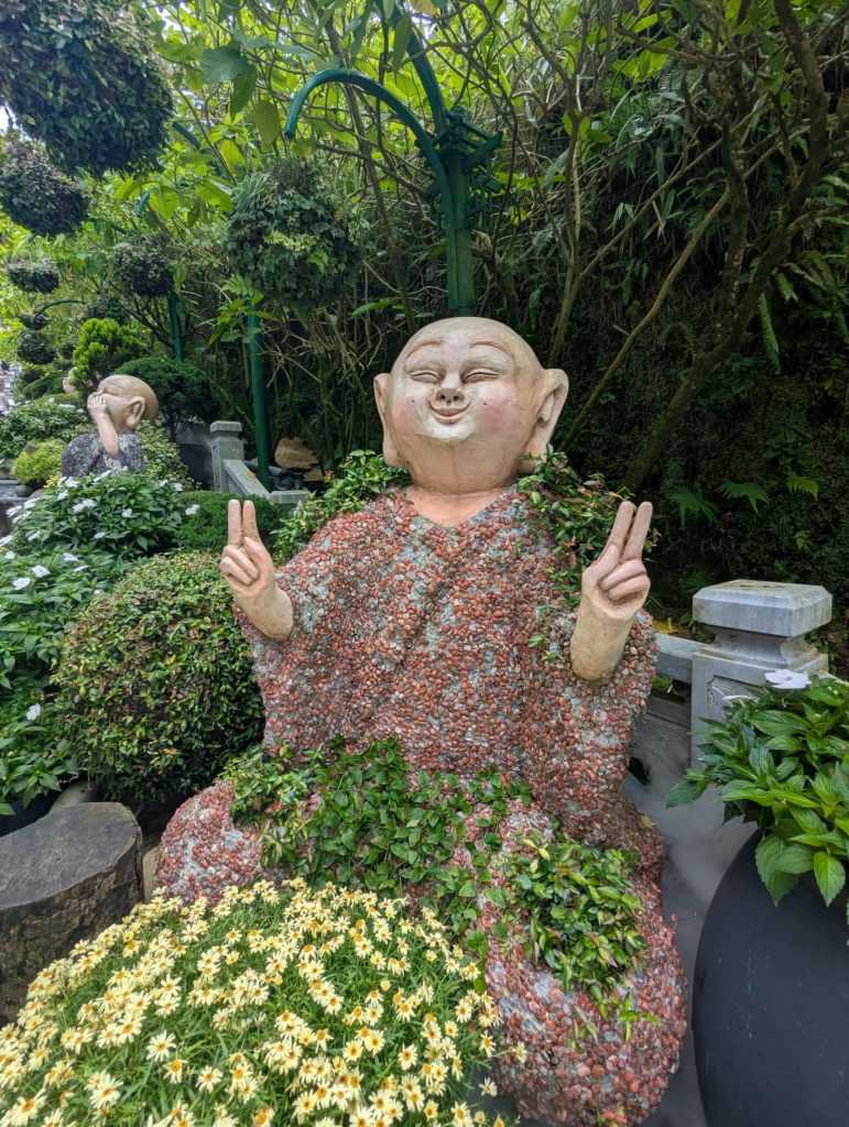 Statue d’un moine souriant faisant un signe de victoire, entourée de végétation à Ba Na Hills.
